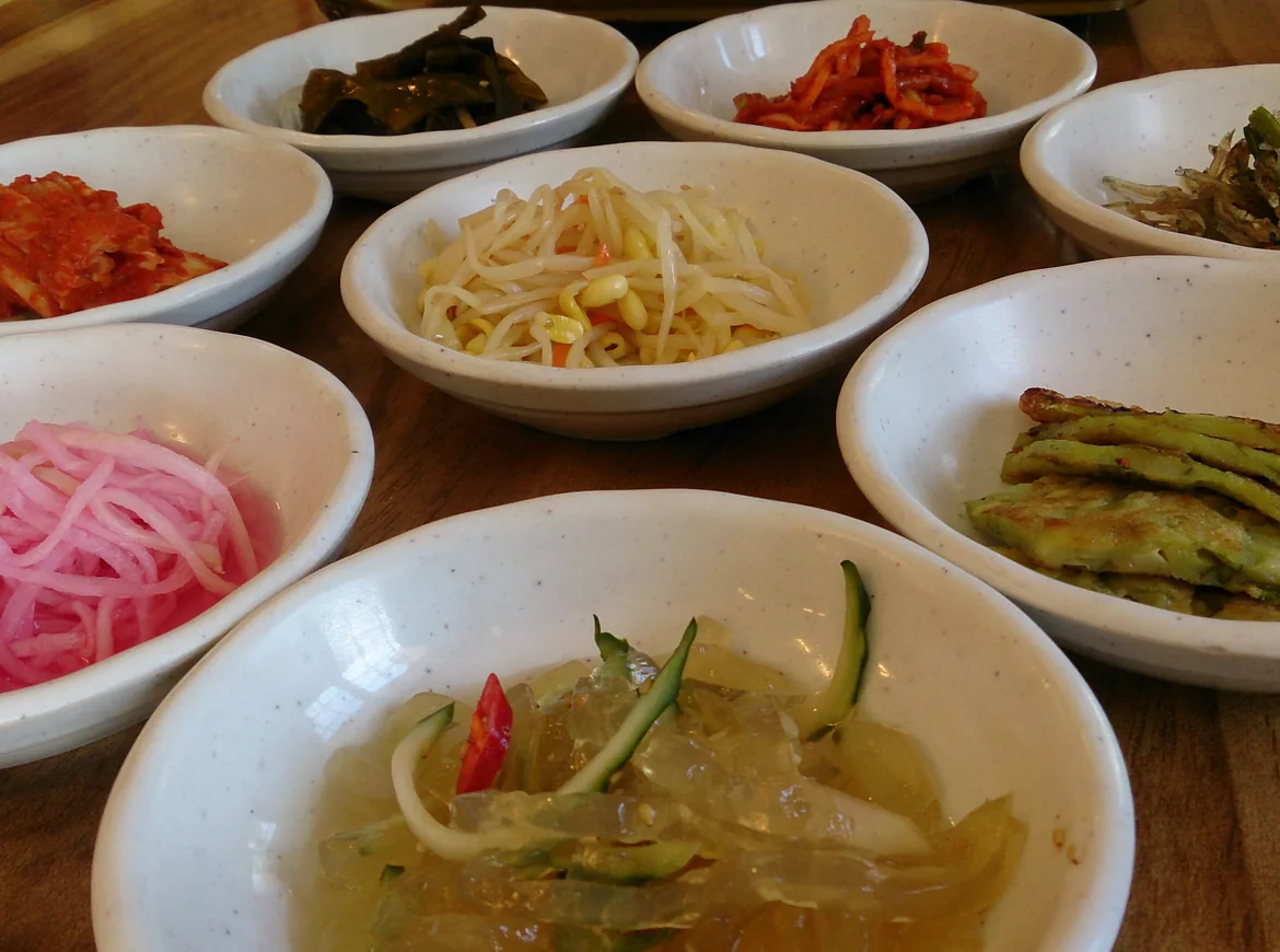 나무 테이블 위 하얀 반찬 그릇들, 그중에는 붉은 김치, 콩나물, 분홍색 단무지, 그리고 맑고 차가운 육수에 담긴 오이가 있다