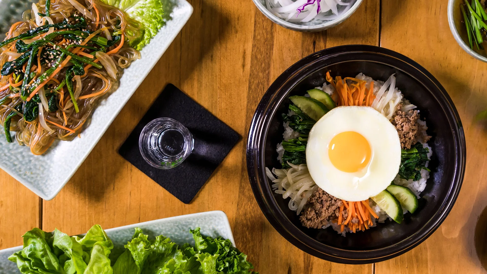 Vista cenital de un cuenco de piedra negra de bibimbap coronado con un huevo frito, zanahoria, espinacas, brotes de soja y carne picada de res sobre una mesa de madera, con un plato de fideos de cristal japchae al lado